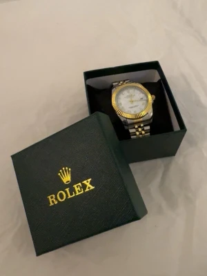 klocka i guld och silver - Snygg klocka med tvåfärgad länk i guld och silver. Urtavlan är vit med romerska siffror och gulddetaljer. Klockan har klassisk rund form och levereras i original ask med logga. Perfekt accessoar för dig som gillar exklusiv stil.