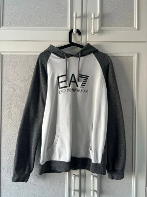 EA7 Emporio Armani hoodie grå/vit - Snygg hoodie från EA7 Emporio Armani i grått och vitt med stor logga på bröstet. Hoodien har huva med dragsko, raglanärmar och en stor magficka. Perfekt för en chill och sportig look.