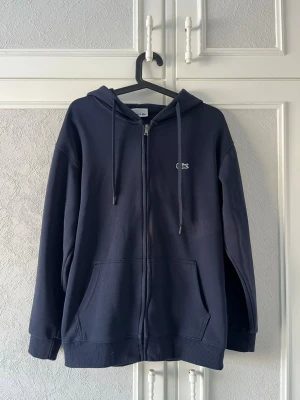 Mörkblå zip-hoodie från Lacoste - Säljer en mörkblå hoodie från Lacoste med dragkedja, huva och snören. Klassisk krokodillogo på bröstet och stora fickor framtill. Perfekt för dig som gillar stilren design och komfort. Materialet är mjukt och känns skönt mot huden.