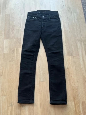 Svarta jeans från Nudie Jeans - Snygga svarta jeans från Nudie Jeans med raka ben. Jeansen har svarta sömmar och diskret logga på bakfickan. Tillverkade i mjuk bomullsdenim som sitter skönt hela dagen.
