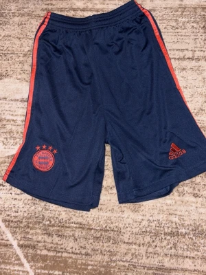 Blå Bayern München shorts Adidas - Mörkblå träningsshorts från Adidas med röda detaljer och Bayern München-logga på ena benet. Shortsen har elastisk midja och röda ränder längs sidorna. Perfekta för fotboll eller träning. Tillverkade i lätt och snabbtorkande material.