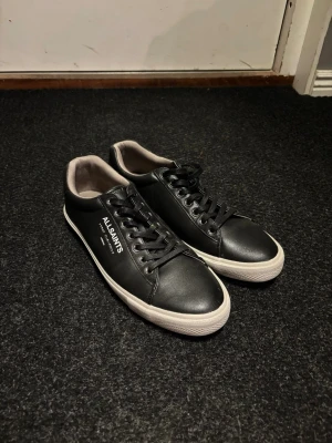 AllSaints - Stilrena svarta sneakers från AllSaints i slätt läder med vit sula och svart snörning. Diskret logga på sidan och klassisk låg modell som passar till många olika outfits. Insidan är ljus och skon har en minimalistisk design.