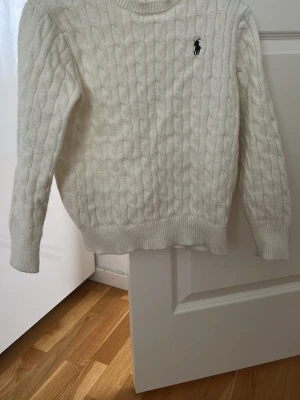 Vit kabelstickad tröja Polo Ralph Lauren - Kabelstickad vit tröja från Polo Ralph Lauren med rund halsringning och svart broderad logga på bröstet. Tröjan har ribbade muddar vid ärmslut och nederkant. Perfekt för lager på lager och klassisk preppy stil.