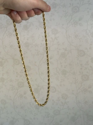 Guldfärgat tvinnat halsband - Snyggt guldfärgat halsband med tvinnad kedja som ger en cool och trendig look. Halsbandet har en klassisk design och passar perfekt för att lyfta din outfit. Materialet ser ut att vara metall med en glansig finish. Det är 18 karat äkta guld planterad i rostfri färg. Låset har varit i grå färg sen jag köpte det så den håller absolut inte på att tappa färg. 