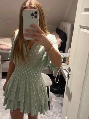 Ljusgrön prickig omlottklänning - Supersöt ljusgrön kortklänning med vita prickar och omlottmodell. Klänningen har volangkant, smock i midjan och korta ärmar. Perfekt för sommarens alla tillfällen och ger en fräsch och trendig look.