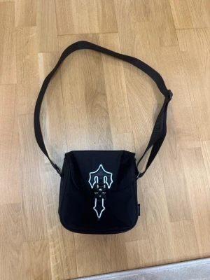 Svart axelväska från Trapstar - Snygg svart axelväska från Trapstar med justerbar axelrem och broderad logga på remmen. Väskan har ett coolt vitt tryck på locket och stängs med spänne. Perfekt för att bära det viktigaste och ger en streetwear vibe.