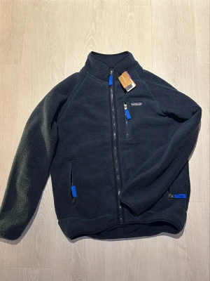 Mörkblå fleecejacka från Patagonia - Mysig mörkblå fleecejacka från Patagonia med hel dragkedja, hög krage och tre fickor med blå detaljer på dragkedjorna. Tillverkad i mjuk fleece som håller värmen och har en sportig look. Perfekt för lager-på-lager och outdoor.