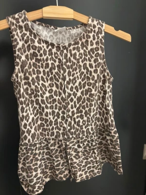 Leopardmönstrad ärmlös topp - Säljer en ärmlös topp med leopardmönster i beige och bruna toner. Toppen har rund halsringning och en lös passform med volang nedtill. Perfekt för dig som vill sticka ut lite extra med ett snyggt leopardmönster