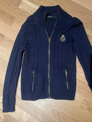 Marinblå kabelstickad ziptröja Lauren stl S - Kabelstickad marinblå tröja från Lauren Ralph Lauren med hel dragkedja, två fickor med guldfärgade dragkedjor och broderad RL-logga på bröstet. Hög krage och ribbade muddar. Perfekt för lager på lager och snygg till jeans.