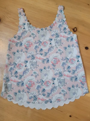 Blommig pastellfärgad linnetopp - Supersöt linnetopp i pastellrosa med blommigt mönster i vitt, blått och grått. Toppen har vågiga kanter både vid axelbanden och nederkanten för en extra gullig look. Perfekt för varma dagar och enkel att styla med jeans eller kjol.