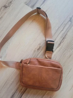 Brun magväska i konstläder - Snygg brun magväska i konstläder med justerbart beige band och svart plastspänne. Väskan har ett huvudfack med dragkedja och en extra ficka framtill. Perfekt för att bära det viktigaste när du är på språng.