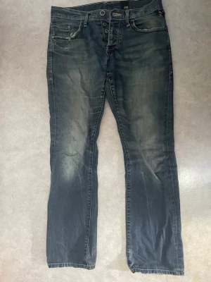 Mörkblå jeans med slitningar från Jack & Jones - Säljer ett par mörkblå jeans från G-Star RAW med snygga slitningar och tvättade detaljer. Jeansen har raka ben, fem fickor och dubbla knappar i midjan. Materialet är kraftigt denim som ger en cool och avslappnad look. Pris kan diskuteras