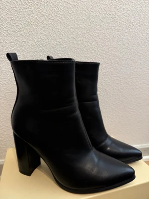 Svarta boots med klack och spetsig tå - Snygga svarta stövletter i slätt skinnimitation med hög blockklack och spetsig tå. Klassisk siluett med dragkedja på insidan och diskret ögla bak. Perfekta för dig som vill ha en stilren och trendig look. 🤗