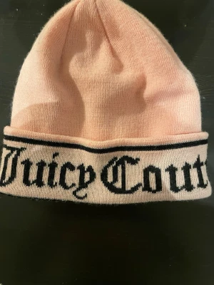Ljusrosa mössa från Juicy Couture - Snygg ljusrosa mössa från Juicy Couture med svart logotyp i gotisk stil runt uppviket. Mjuk och stretchig, perfekt för att hålla värmen och ge en trendig touch till din outfit.