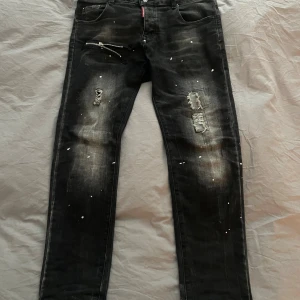 Svarta  jeans från Dsquared2 - Säljer ett par svarta jeans från Dsquared2 med slitningar, färgstänk och dragkedjedetalj vid fickan. Jeansen har en slim fit passform och coola destroyed detaljer på båda benen. Perfekta för dig som gillar streetwear och edgy stil.