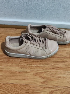 Beige Fred Perry sneakers - Snygga beige sneakers från Fred Perry med klassisk logga på sidan. Skorna har lågt skaft, matchande beige snörning och en vit sula. Materialet är en mix av mocka och glansigt skinn på tån för en stilren look. Perfekta för dig som gillar enkel och clean stil.