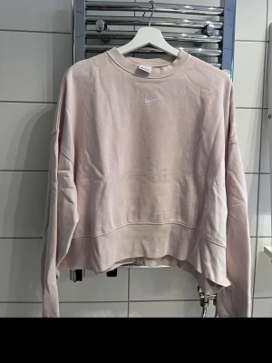 Beige cropped hoodie från Nike - Säljer en beige cropped hoodie från Nike med broderad vit Swoosh-logga på bröstet. Hoodien har rund halsringning, långa ärmar och är gjord i mjukt bomullsmaterial. Perfekt för en avslappnad och trendig look.