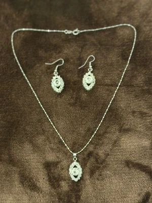 Silverfärgat smyckeset med örhängen 925S - Snyggt smyckeset i silverfärg med halsband och matchande örhängen. Både halsbandet och örhängena har ovala hängen med gnistrande stenar och detaljerad kant. Perfekt för dig som gillar unika accessoarer med lite extra bling.