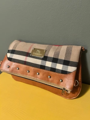 Rymlig ny axelväska klassiska rutmönstert  - Ny Snygg axelväska som liknar Burberry i klassiskt beige, svart och rött rutmönster. Väskan har detaljer i brunt konstläder, silverfärgade nitar och dragkedja samt en metallbricka med logga framtill. Justerbar axelrem flyttbar så du kan bära väskan så du får ner din platta och kalender eller med ett övervikande lock som Halverar det rymliga huvudfacket 