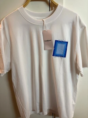 Vit t-shirt från Casablanca - Vit t-shirt från Casablanca med tryck i blå toner både fram och bak. Grafiskt motiv med texten 'Casa Blanca' i vitt ovanpå blå rektanglar. Klassisk passform och rund halsringning. Perfekt för dig som gillar stilrena och moderna plagg.