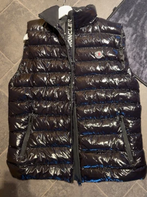  dunväst från Moncler - Snygg svart dunväst från Moncler med glansigt yttermaterial och klassisk Moncler-logga på bröstet. Västen har två dragkedjeförsedda fickor framtill och hög krage med Moncler-tryck på insidan. Perfekt för lager-på-lager och streetstyle.(inte äkta! vet inte vart ifrån ,fick den)