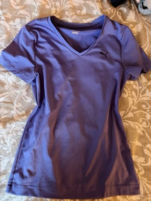 Lila v-ringad t-shirt från Puma - Snygg lila t-shirt från Puma med v-ringning och diskret Puma-logga på bröstet. T-shirten har korta ärmar och är gjord i mjuk bomull, perfekt för en avslappnad och sportig stil.