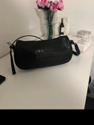 Svart axelremsväska i krokodilmönster - Snygg svart handväska med axelrem och trendigt krokodilmönster. Väskan har en avlång form och dragkedja upptill. Perfekt accessoar för att lyfta din outfit med en edgy touch.