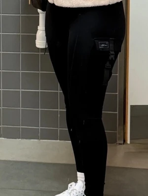 Svarta cargobyxor från I Can I Will - Säljer ett par svarta cargobyxor från I Can I Will med hög midja och smal passform. Byxorna har praktiska fickor på sidorna, dragkedjedetalj och en patch med logga på benet. Perfekta för en sportig och avslappnad stil.