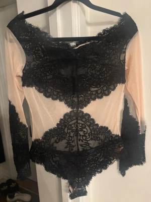  Juniorrosa body med svart spets - Säljer en snygg body i ljusrosa mesh med detaljer i svart spets. Bodyn har långa ärmar och off-shoulder design. Spetsen är placerad över byst, midja och ärmslut för en elegant look. Materialet är genomskinligt mesh och spets, vilket ger en lyxig känsla sopassad för trendiga outfits.