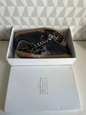 Maison margiela - Unika svarta sneakers med färgstänk i vitt, rött, blått och gult. Skorna har vita snören, låg profil och en klassisk gummisula. Ovandelen är i läder och designen ger en konstnärlig och modern vibe. Perfekta för dig som vill sticka ut med din stil.