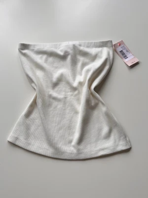 Vit ribbad tubtopp med slits - Trendig vit ribbad tubtopp. Toppen är axelbandslös och har en liten slits på sidan för extra detalj. Perfekt att styla med höga jeans eller kjol för en clean look. Helt oanvänd med prislapp på