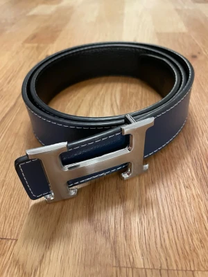 H bälte - Snyggt blått läderbälte med ett stort, silverfärgat H-spänne framtill. Bältet har vita kontrastsömmar längs kanterna och en klassisk, stilren design som passar till jeans eller kostymbyxor.