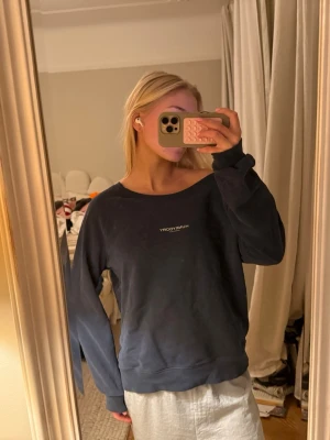 Mörkblå sweatshirt från Hunkydory - Mörkblå sweatshirt från Hunkydory med vit logotyp på bröstet. Tröjan har en avslappnad passform, rund halsringning och långa ärmar. Perfekt för en chill look och enkel att matcha med jeans eller mjukisbyxor.
