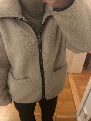 Beige teddyjacka med dragkedja - Mysig beige teddyjacka från ARKET med hög krage och hel dragkedja framtill. Jackan har två stora fickor på sidorna och är gjord i fluffigt teddy-material. Några tecken på användning så som att den har blivit lite nopprig inuti (se bilderna) men inget som syns när man bär jackan!! Annars väldigt fint skick. Denna modell säljs inte längre men nypris var runt 1700kr