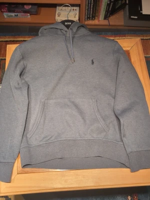 Blå hoodie från Polo Ralph Lauren - Snygg grå hoodie från Polo Ralph Lauren med klassisk broderad logga på bröstet. Hoodien har huva med dragsko, magficka och ribbade muddar vid ärmar och nederkant. Tillverkad i mjukt bomullsmaterial som är perfekt för en avslappnad stil.