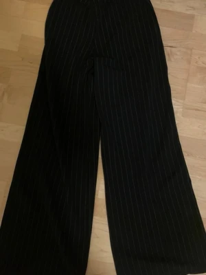 Svarta vida kostymbyxor med ränder - Säljer ett par svarta kostymbyxor med vita tunna kritstreck. Byxorna har hög midja och vida ben för en avslappnad men ändå dressad look. 