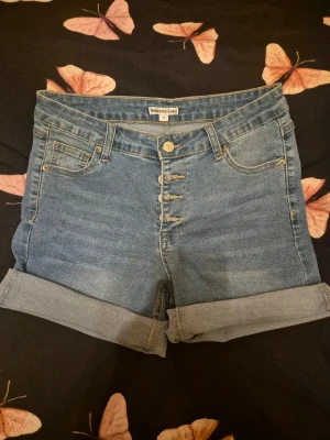 Blå jeansshorts från Sincerely Love - Säljer ett par klassiska blå jeansshorts från Sincerely Love med hög midja och uppvikta benslut. Har fem fickor och stängs med fyra synliga knappar framtill. Perfekta till sommaren och lätta att matcha med allt.
