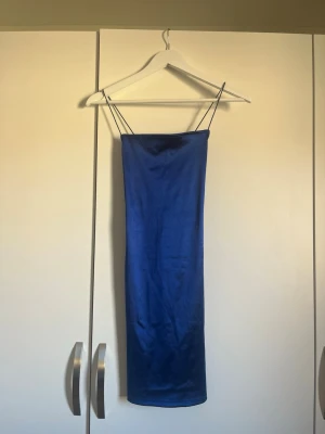 Blå satin klänning med smala band - Säljer en stilren blå klänning i satin med tunna axelband och rak passform. Klänningen är kort och har en enkel, elegant design som ger en lyxig känsla. Perfekt för dig som gillar minimalistisk stil och vill ha något som sticker ut.