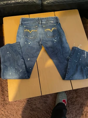 Blå jeans med gult broderi - Säljer ett par blå jeans från Wrangler med coolt gult broderi på bakfickorna och slitna detaljer samt vita färgstänk. Klassisk femficksmodell och rak passform. Perfekta för en avslappnad och trendig look.