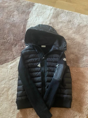 Svart dunväst med huva från Moncler - Snygg svart dunväst från Moncler med stickade ärmar och stor huva. Västen har dragkedja framtill, Moncler-logga på ärmen och quiltad design. Perfekt för lager-på-lager och streetwear-stil. Materialet är en mix av dun och stickat tyg.