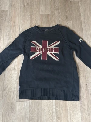 Svart stickad tröja Morris S - Svart stickad tröja från Morris med Union Jack-motiv i vinrött och beige på bröstet. Rund halsringning och ribbade muddar. Perfekt för dig som gillar klassisk stil med en twist. Märkesdetaljer på ärmen och insidan. Vid snabb affär kan pris sänkas.