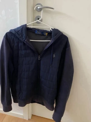 Mörkblå Polo Ralph Lauren cardigan - Snygg mörkblå hoodiejacka från Polo Ralph Lauren med quiltad front, huva och dragkedja. Jackan har en broderad logga på bröstet och ribbade muddar. Perfekt för lager på lager och chill vardagsstil. Storlek M-175.