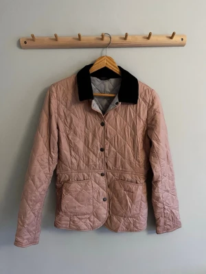 Quiltad rosa jacka med svart krage - Quiltad jacka från barbour i ljusrosa med svart krage. Jackan har knappar framtill, två stora fickor och en klassisk passform. Perfekt för vår och höst när du vill ha en stilren och bekväm look.