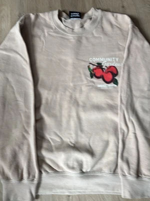 Beige sweatshirt med tryck från ADANOLA - Säljer en beige sweatshirt med rund hals och tryck av röda tomater samt texten 'COMMUNITY GARDEN' på bröstet. Tröjan har ribbade muddar vid ärmslut och nederkant. Perfekt för en avslappnad och trendig look.