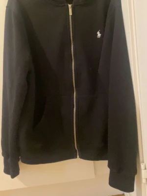 Svart hoodie från Polo Ralph Lauren - Klassisk svart hoodie med dragkedja från Polo Ralph Lauren. Tröjan har huva, två fickor framtill och den ikoniska vita logon broderad på bröstet. Tillverkad i mjukt material som passar perfekt till en avslappnad stil. 