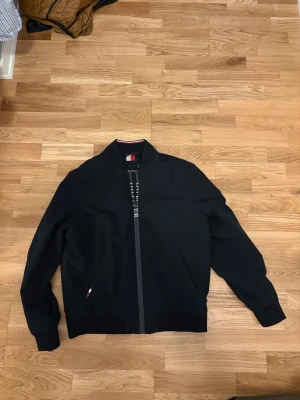 Svart bomberjacka Tommy Hilfiger - Stilren svart bomberjacka från Tommy Hilfiger med dragkedja framtill och diskreta fickor på sidorna. Jackan har klassisk bomberkrage och muddar vid ärmslut och midja. Detaljer med Hilfiger-logga på insidan av dragkedjan och liten flaggdetalj vid fickan. Vänstra fickan går inte att stänga pågrund av att en knapparna har ramlat av men inga håll inuti fickan.