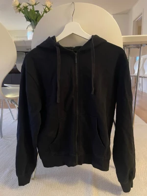 Svart zip-hoodie med fickor - En klassisk svart hoodie med dragkedja framtill och två stora fickor. Modellen har huva med dragsko och ribbade muddar vid ärmar och midja. Perfekt för en avslappnad och stilren look.