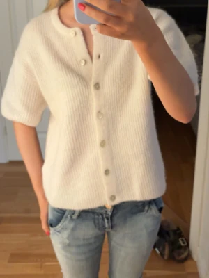 Vit kortärmad kofta med knappar - Mysig vit kofta med korta ärmar och rund hals. Koftan har ribbad struktur och stängs med pärlemorskimrande knappar framtill. Perfekt att slänga över en topp för en enkel och clean look.