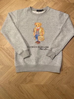 Grå sweatshirt Polo Bear Ralph Lauren - Säljer en grå sweatshirt från Polo Ralph Lauren med det ikoniska Polo Bear-motivet på bröstet. Tröjan har rund hals, ribbade muddar och nederkant samt mjukt fleecefoder på insidan. Perfekt för dig som gillar klassisk streetwear med en twist. Bara att höra av sig vid frågor☺️ Kan kanske gå ner i pris lite men inte något extremt eftersom priset redan är ganska lågt!