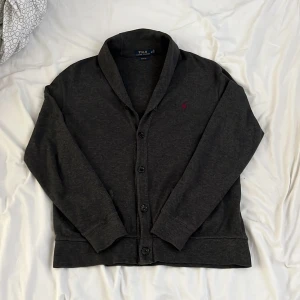 Ralph Lauren cardigan - Säljer en stilren mörkgrå kofta från Polo Ralph Lauren med knappar framtill och en liten broderad logga i rött på bröstet. Perfekt för en avslappnad men ändå snygg look.